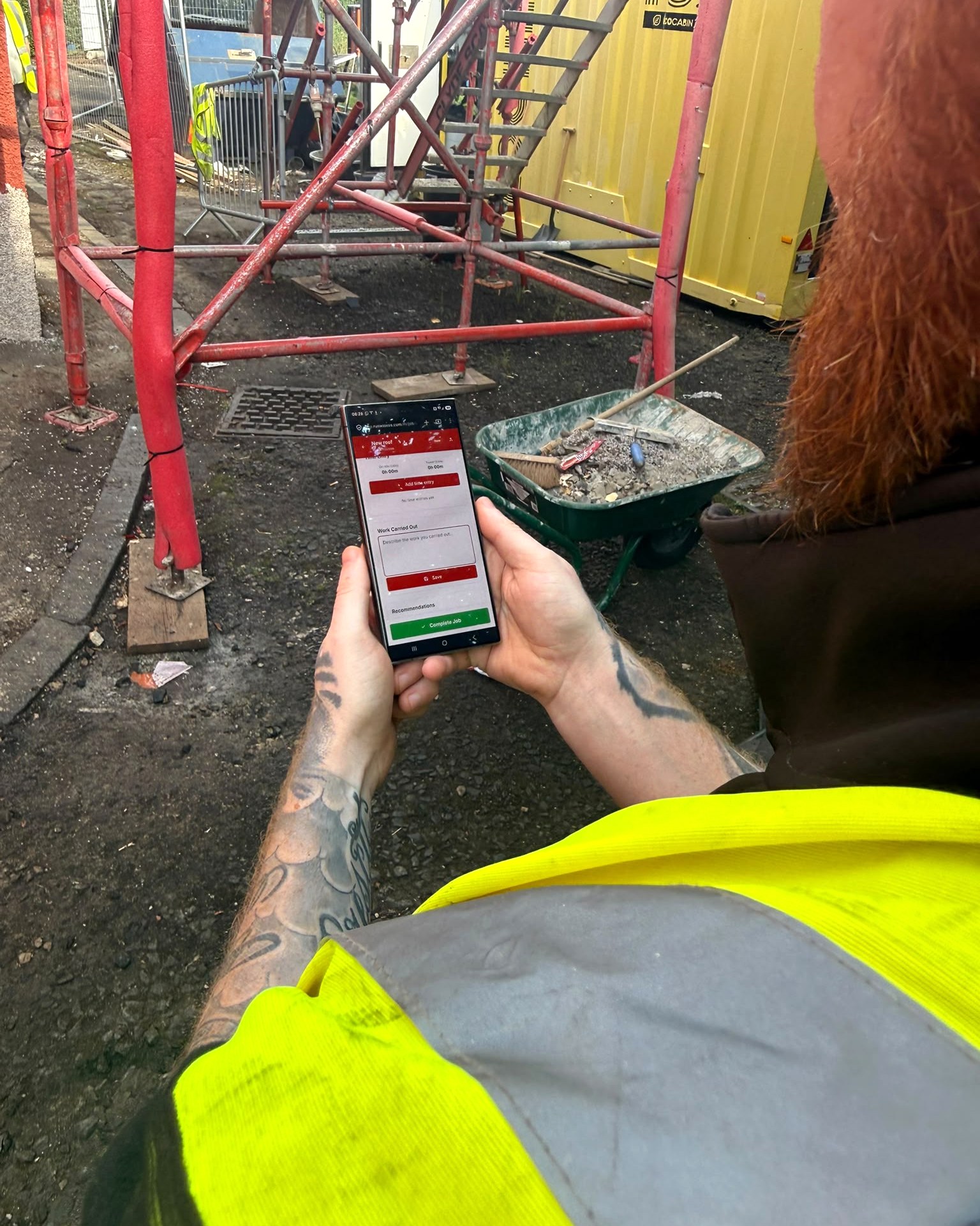 Contractor filling out a RunwiseOS digital job sheet on his phone on a UK building site, wheelbarrow and scaffolding around him